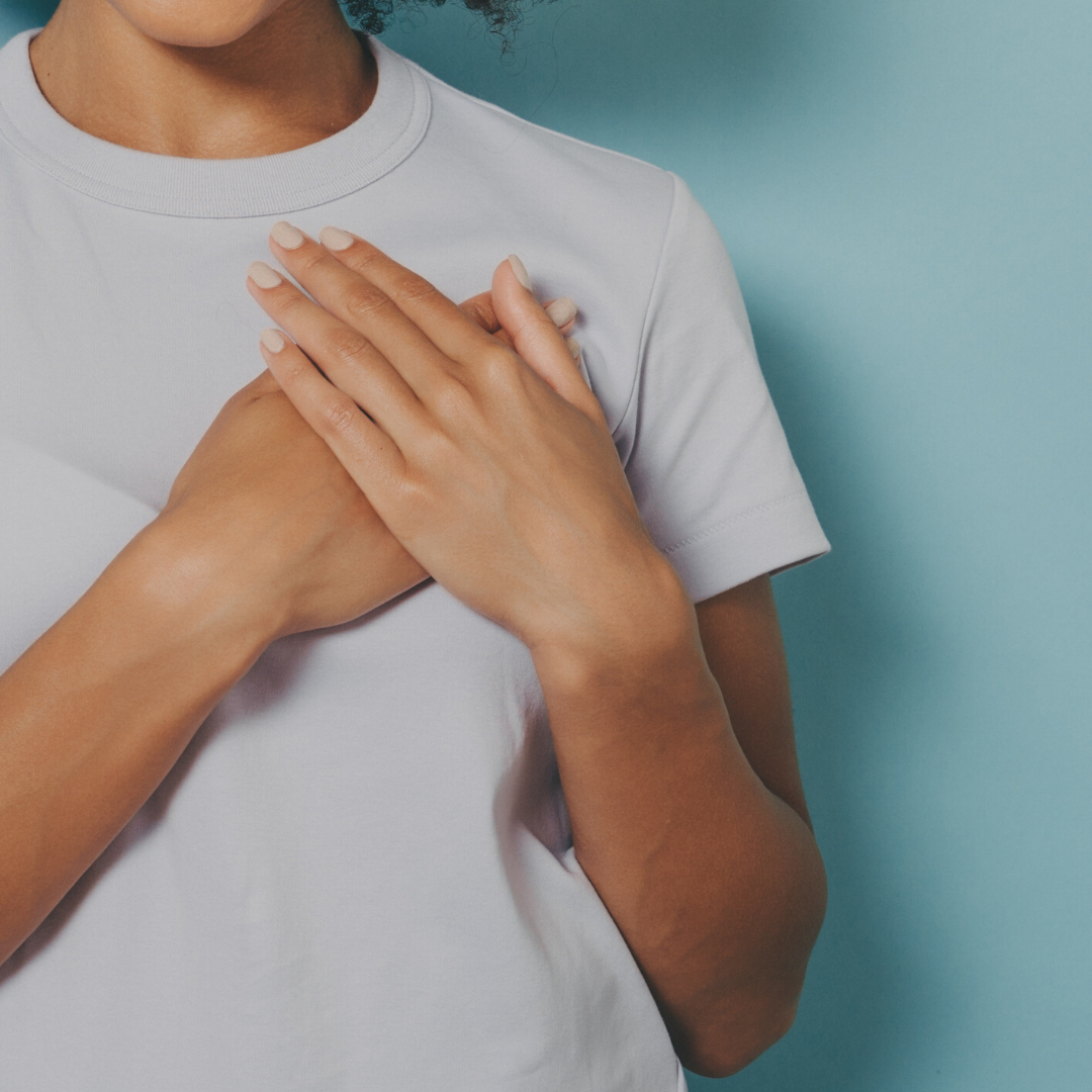 Woman in white t-shirt holding her hands over her heart (image via Canva)